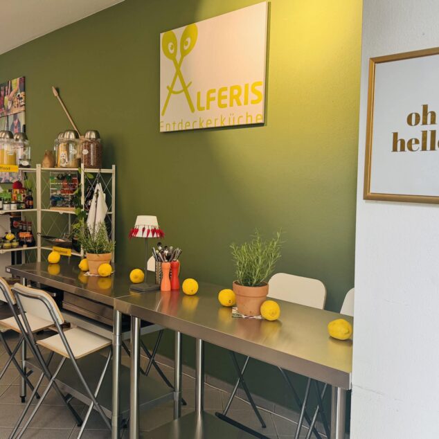 Innenraum der Alferis Entdeckerküche in Tutzing mit Edelstahltisch, frischen Zitronen, Kräutertöpfen und dem Markenlogo an der olivgrünen Wand